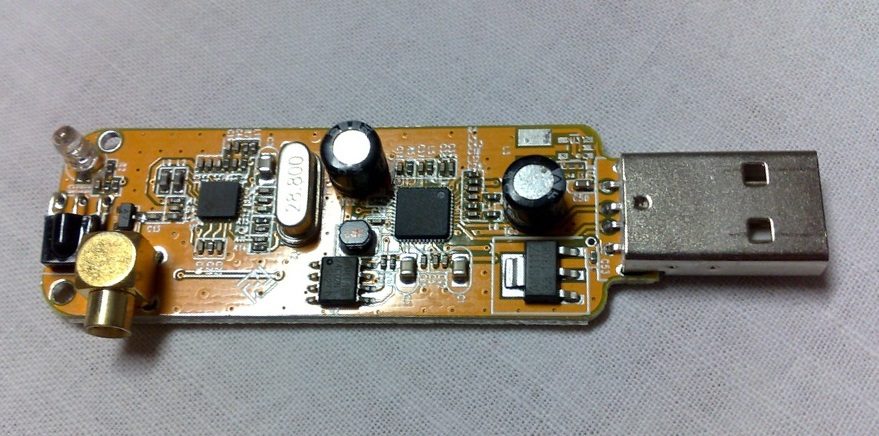 The bare circuit board of a cheap USB TV stick: a small square tuner chip on the left, a silver crystal marked 28.800, a larger square Realtek chip, capacitors, an antenna socket at one end and a USB plug at the other