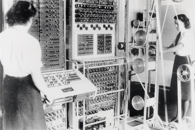 Two women operating Colossus, a wall of valves and switches, with a paper tape running over pulleys on a frame