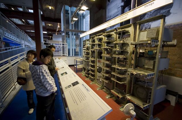 A long row of grey equipment racks in a museum gallery, with visitors looking at them