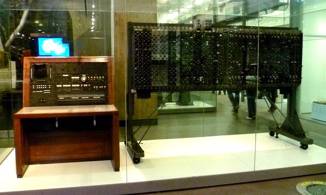 A wooden control desk beside a tall cabinet of valves behind glass in a museum