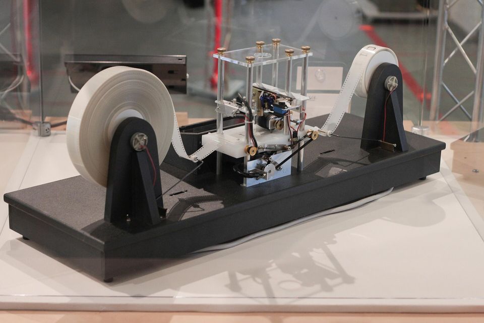 A small working Turing machine on a display stand: a tape wound between two white reels, passing through a clear box that holds the read and write head