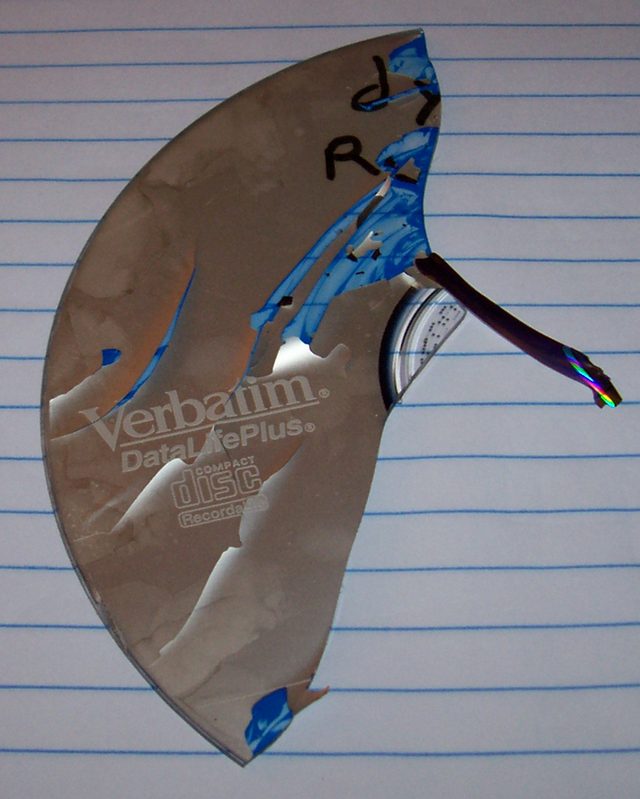 A large wedge-shaped piece of a broken CD-R lying on lined paper, its silver label side partly peeled away in patches, with a sliver of disc still attached